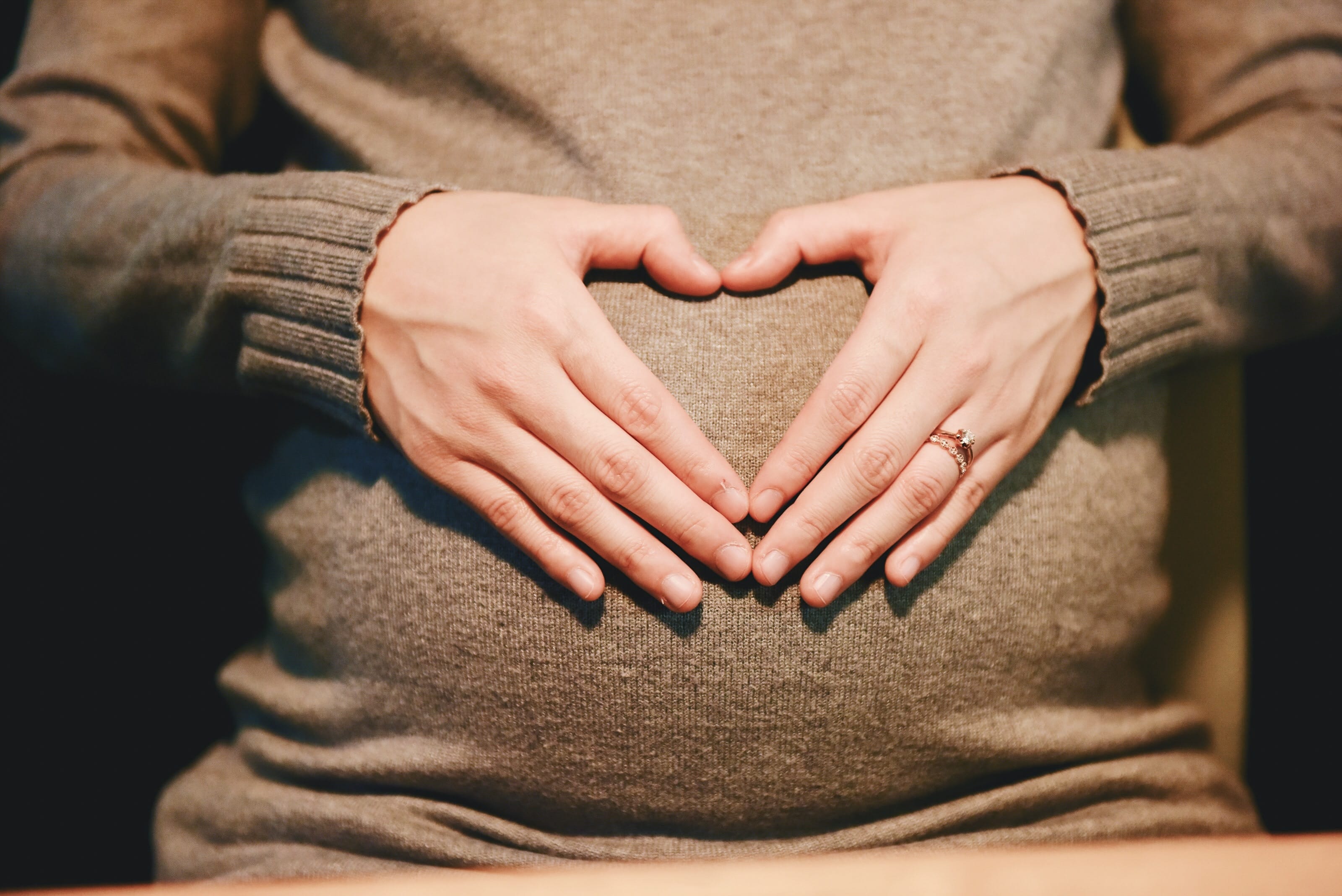 A pregnant woman in a sweater forming a heart with her hands on her belly