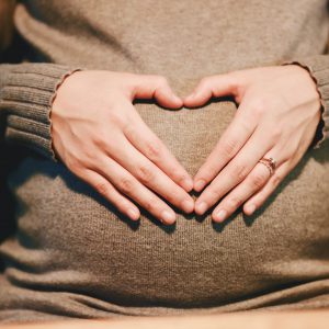 A pregnant woman in a sweater forming a heart with her hands on her belly